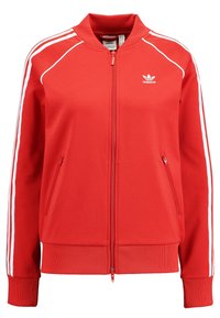 Červená zipová bunda vyrobená z hladkého materiálu, se zelenými bočními pruhy, žebrovanými manžetami a dvěma předními kapsami. Na hrudi je logo Adidas.