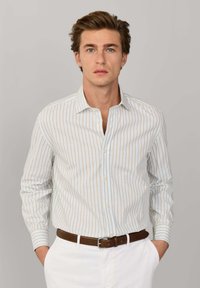 Junger Mann mit blauen Augen, der ein beige-blau gestreiftes Hemd trägt, das in weiße Hosen mit braunem Gürtel gesteckt ist, vor einem grauen Hintergrund.