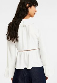 Hvid bluse med lange ærmer og flæsedetalje ved halsen, struktureret stof, elastisk talje og udvidede manchetter, parret med mørke denimbukser.