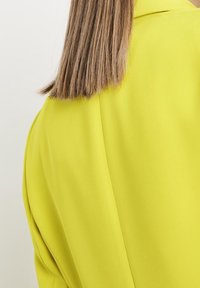 Blusa gialla su misura con texture liscia, dotata di un colletto strutturato e dettagli drappeggiati sulla spalla.