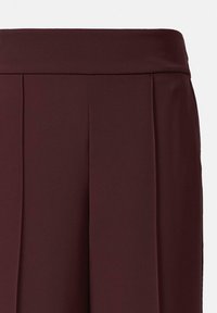 Burgunderfarbene, plissierte Hose mit breite Bund. Glatte, matte Textur ohne sichtbare Beschläge oder Muster. Fokus auf den weichen Fall des Stoffes.