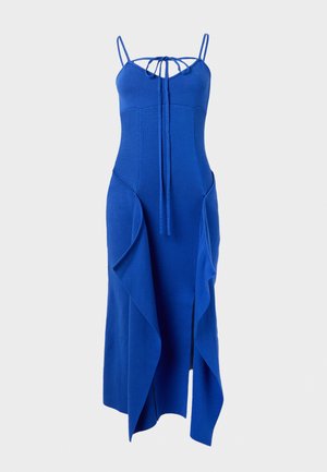 Femme portant une robe midi bleue sans manches avec un détail noué à l’avant et des panneaux drapés asymétriques, associée à des sandales noires à talons.