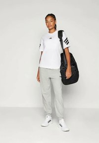 T-shirt branca de manga curta da Adidas com riscas pretas, calças de fato cinzentas ajustadas e mochila preta. Modelo a usar sapatilhas brancas da Adidas.