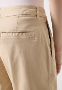 Gros plan d’un pantalon beige montrant une poche arrière et un logo subtil "BOSS" sur le côté droit près de la ceinture.