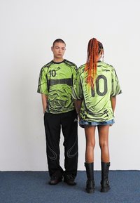 Grønne grafiske t-skjorter med store tall "10" iført svarte bukser og et denim skjørt. Den kvinnelige figuren kombinerer skjorten sin med svarte støvler.