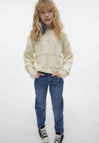 Stickad krämfärgad tröja med öppna mönster, matchad med ljusblå jeans. Svartvita högskurna sneakers fulländar outfiten.