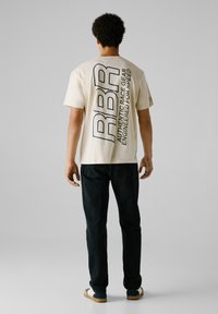 Camiseta beige con un gran gráfico en texto azul oscuro en la parte posterior, que dice "RBR AUTHENTIC RACE GEAR ENGINEERED FOR SPEED". Jeans negros y zapatos casuales.