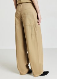 Frau in weiten, locker sitzenden khakifarbenen Hosen mit großen Rück- und Taschen an der Seite, stehend in schwarzen spitz zulaufenden Schuhen vor einem schlichten Hintergrund.