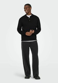 Melns polo džemperis ar apkakli un trim pogām, valkāts pāri baltai kreklam. Kombinēts ar vaļīgām tumši pelēkām biksēm un melniem zābakiem.