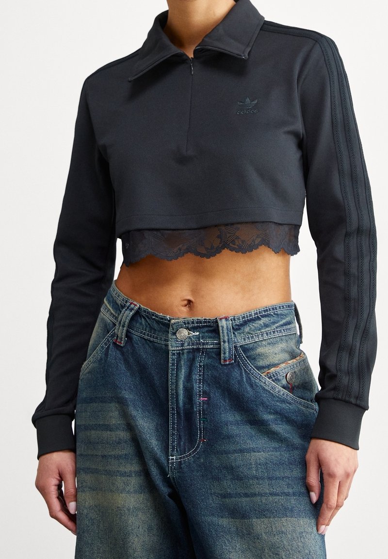 Top negro corto de manga larga Adidas con encaje en el borde, combinado con jeans azules desteñidos de tiro alto y baggy, en una persona de pie.