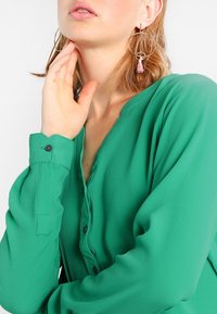 Vihreä smaragdinvärinen napitettava blousi pitkillä hihoilla, pehmeä rakenne ja mustat napit; mallilla on käsi lähellä kasvoja ja korvissa renkaat.
