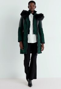 Tumši zaļš piepūsts mētelis ar kažokādas odere kapuci, kuram ir melni ādas akcenti uz piedurknēm, valkāts virs baltas krekla un melnām platām biksēm.