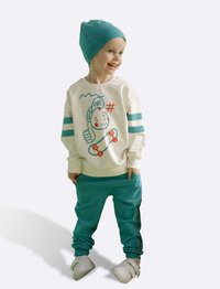 Jeugdoutfit met een crèmekleurige sweatshirt met een cartoonontwerp en teal accenten, gecombineerd met teal joggingsbroek en witte schoenen.