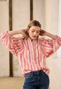 Moteris, pasipuošusi dideliais lankiniais auskarais, dėvi rožinę ir baltą dryžuotą laisvą palaidinę su pūstomis rankovėmis ir mėlynas džinsus, stovinti patalpose.