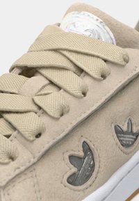 Bézs színű, velúr sportcipő lapos fűzőkkel, texturált panelekkel, és oldalt áttetsző Adidas trefoil logókkal. Fehér gumitalp és varrás.