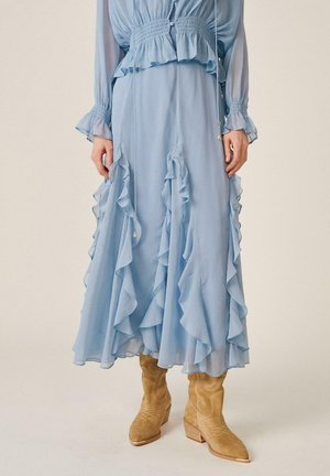 Falda midi azul claro con volantes verticales, acompañada de una blusa a juego y botas cowboy de ante color beige sobre un fondo liso.