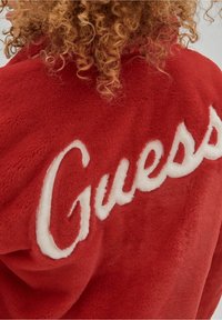 Rode fleecejas met een prominent wit geborduurd "Guess"-logo op de achterkant, met een zachte textuur en warm ontwerp.