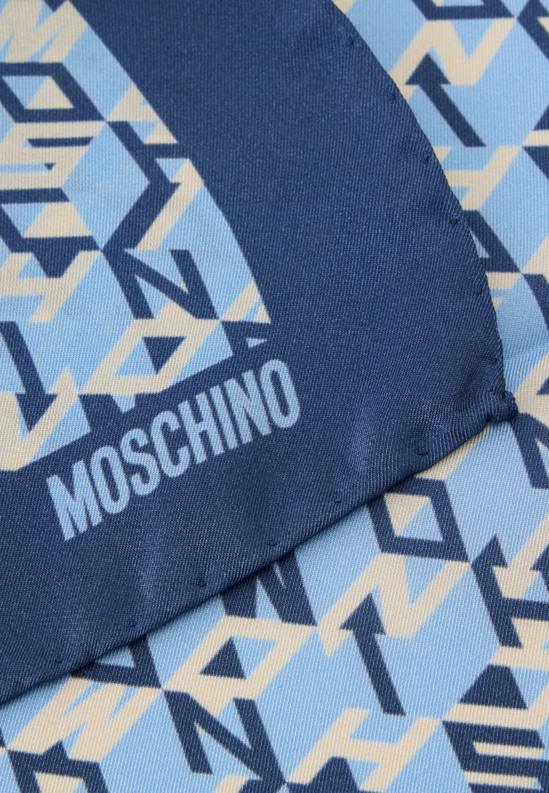 Primer plano de tela doblada con patrón geométrico azul y beige y la palabra "MOSCHINO" impresa en azul claro en la sección azul más oscura.