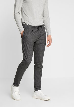 Mann trägt ein graues Langarmshirt, dunkelgraue Jogginghosen mit Kordelzug, weiße Socken und weiße Sneakers und steht vor einem schlichten Hintergrund.