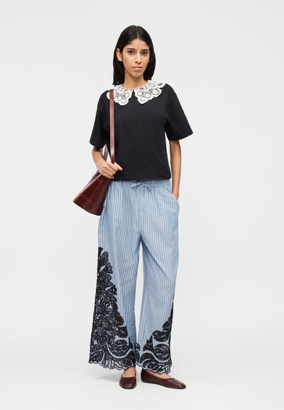 Femme debout portant un haut noir avec un col en dentelle blanche, un pantalon rayé bleu avec des détails en dentelle noire, des chaussures marron et un sac à bandoulière marron.