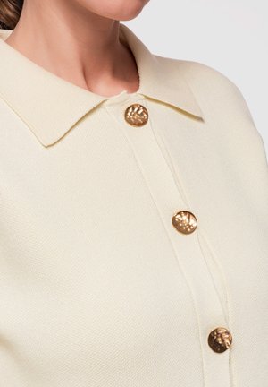 Gros plan d'une femme portant une chemise en tissu texturé crème avec un col pointu et trois boutons dorés martelés brillants sur le devant.