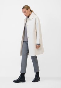 Beige Wollmantel mit strukturiertem Design, ausgestattet mit goldenen Knöpfen, seitlichen Taschen und einem breiten Kragen, getragen über einem grauen Pullover und grauen Hosen.