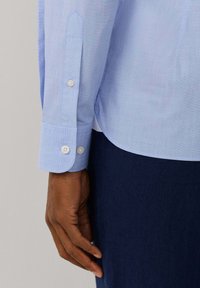 Tmavá ruka odpočívající vedle manžety světle modré košile s dlouhým rukávem se dvěma bílými knoflíky a tmavě modrých kalhot.