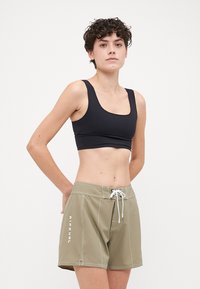 Fekete sportmelltartó, amelyet olajzöld, térd hosszúságú úszónadrággal párosítanak, fehér zsinórral és az oldalon fehér "RIPCURL" felirattal.
