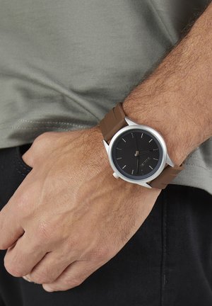 Round black watch face with silver bezel, brown silicone strap, minimalist hour markers, worn on a hand against a gray fabric background.