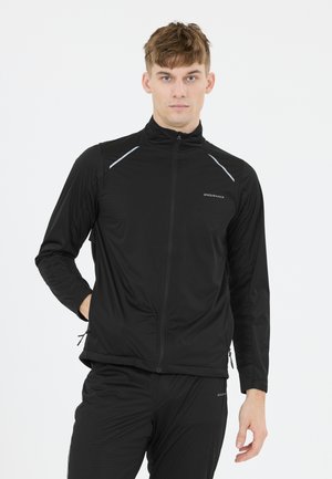 Ung mand iført sort sporty jakke med lynlås og matchende bukser, stående med den ene hånd i lommen mod en ensfarvet hvid baggrund.