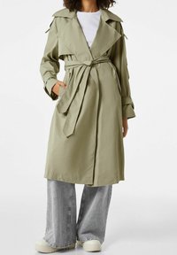 Khaki trench coat s páskem v pase, širokým límcem, dlouhými rukávy a bočními kapsami. Kombinováno se šedými širokými džínami a bílými teniskami.