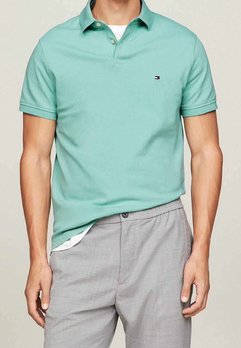 Polo vert menthe avec un col et des manches courtes, arborant un petit logo sur la poitrine. Porté avec un pantalon gris clair.