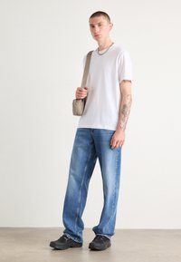 T-shirt blanc à manches courtes, jean bleu ample, baskets noires et sac bandoulière beige. Présente un tatouage sur le bras droit.