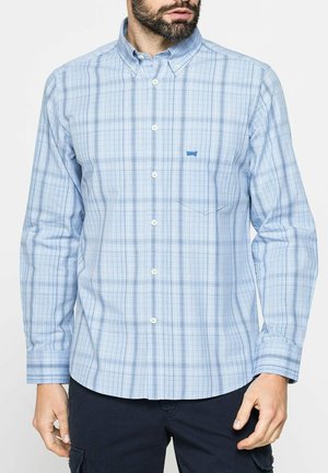 Uomo che indossa una camicia a quadri azzurro chiaro con taschino sul petto e pantaloni scuri, in piedi davanti a uno sfondo neutro.