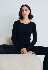 Donna con capelli castani mossi e occhi blu, che indossa un top nero a maniche lunghe e leggings, seduta a gambe incrociate su un divano di tessuto color crema.