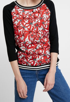 Langærmet top med sorte ærmer og et levende rødt og hvidt blomstermønster på torsoen, båret med blå jeans.