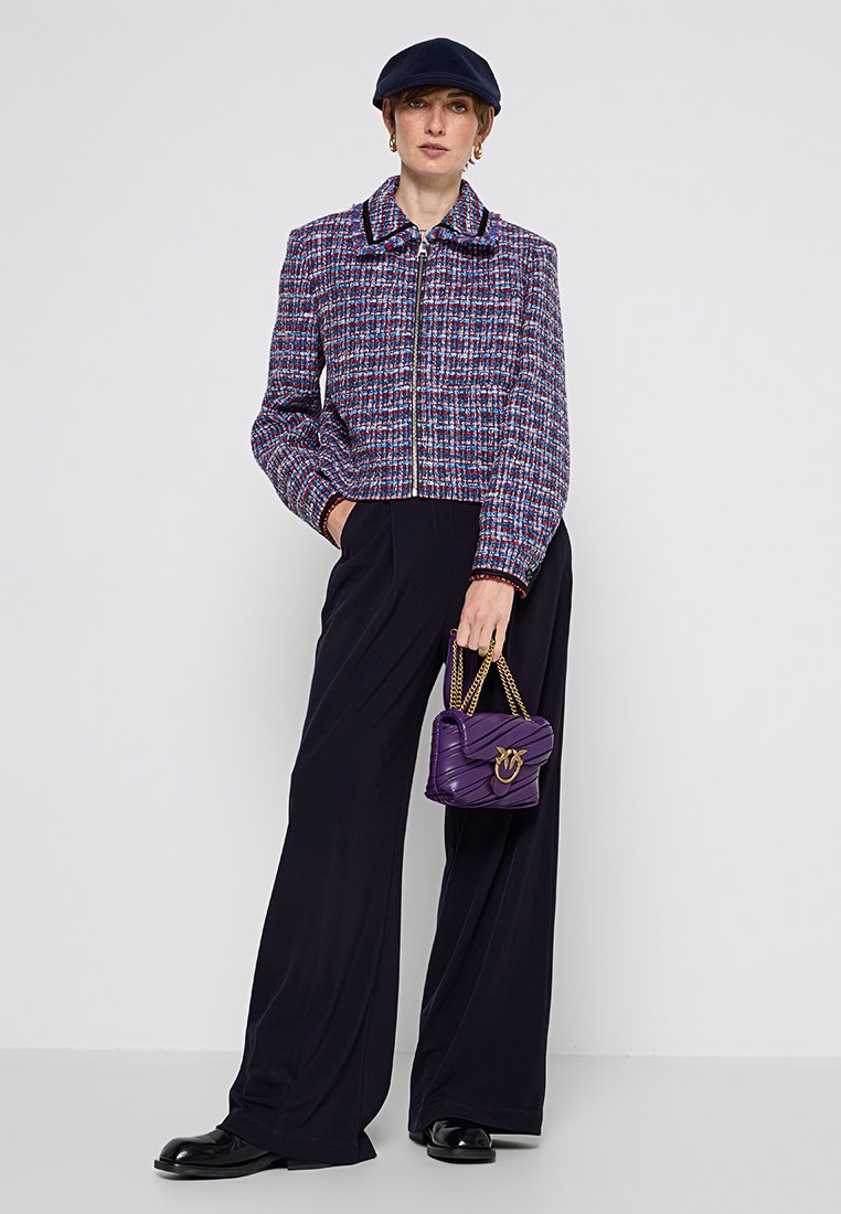 Giacca in tweed tessuto nei colori viola, rosso e blu, con colletto e chiusura a zip, abbinata a pantaloni neri a gamba larga. Piccola borsa viola con dettagli dorati.
