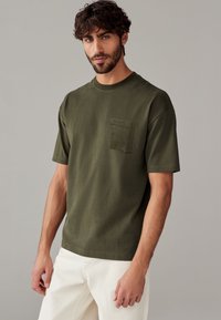T-shirt verde oliva con colletto tondo, maniche corte e taschino sul petto. Il tessuto appare liscio con una vestibilità rilassata.