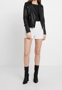 Žena oblečená v černé kožené bundě, černém krátkém topu, bílých krajkových šortkách a černých kotníkových botách na podpatku stojící na bílém pozadí.