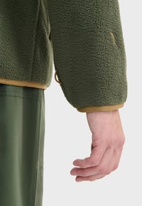 Grøn fleecejakkeærme med en tekstureret overflade, der har en tan elastisk manchet og en justerbar snor. Hånd synlig ved enden.
