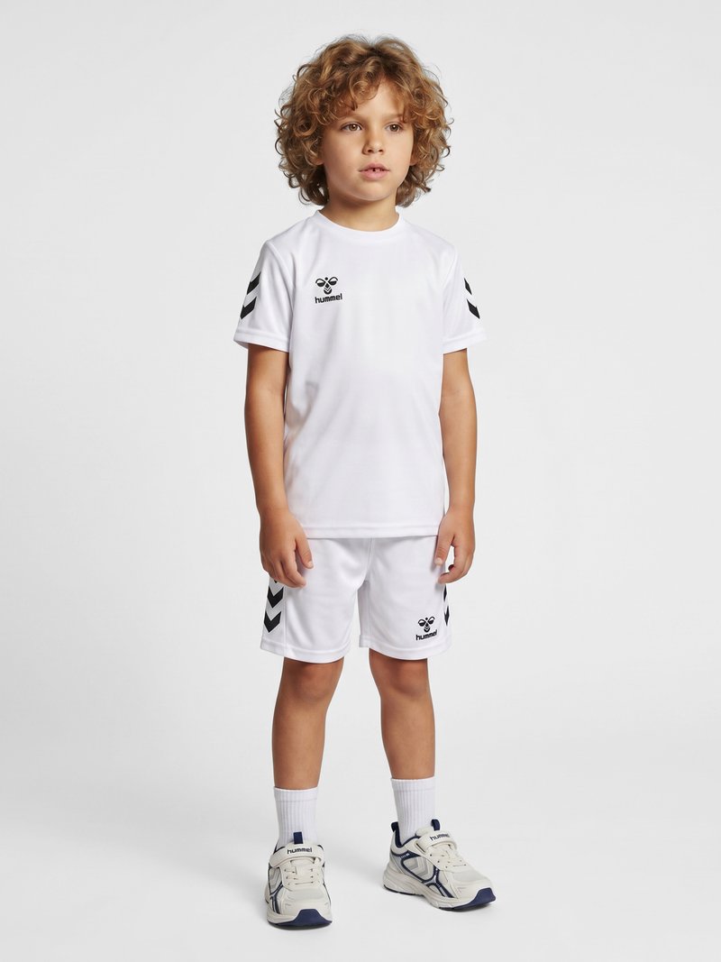 Jonge jongen met krullend haar staat in witte sportkleding met zwarte logo's en witte sneakers tegen een egale lichte achtergrond.