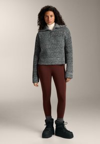 Šedý texturovaný pletený svetr s límečkem a polovičním zipem, v kombinaci s vínovými legínami a černými zimními botami.
