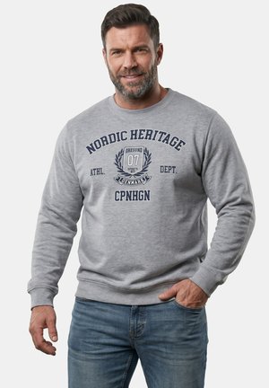 Mand med skæg iført grå "Nordic Heritage" sweatshirt og blå jeans, står mod en lys baggrund med den ene hånd i lommen.