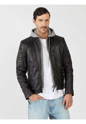 Man draagt een zwarte leren jas met doorgestikte mouwen over een grijze hoodie en wit overhemd, lichtblauwe spijkerbroek, één hand in de zak.