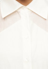 Κοντινό πλάνο ενός λευκού πουκαμίσου με γιακά και κουμπιά στο εμπρόσθιο μέρος, με διαφανές ύφασμα στο πάνω μέρος του στήθους.