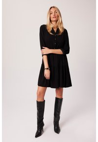 Czarna sukienka do kolan z przodem na guziki, kołnierzykiem i rozkloszowanym dołem. Stylizowana z wysokimi czarnymi botkami. Materiał wydaje się gładki i miękki.