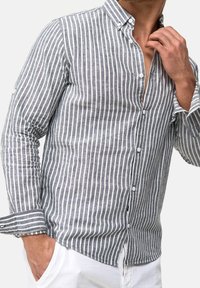 Man draagt een grijs-wit verticaal gestreept overhemd met knopen en witte broek, met één hand in de zak en de andere tegen de kraag.