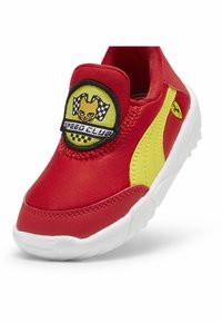 Piros és sárga slip-on sportcipő, gumi talppal, kerek fekete és sárga logóval, sima textil felsőrésszel.