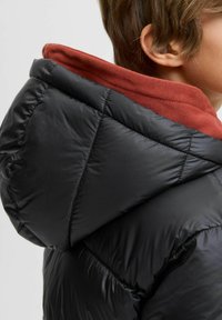 Doudoune noire avec un col intérieur rouge distinctif, arborant une texture matelassée et une capuche, mettant en avant un design élégant et moderne.