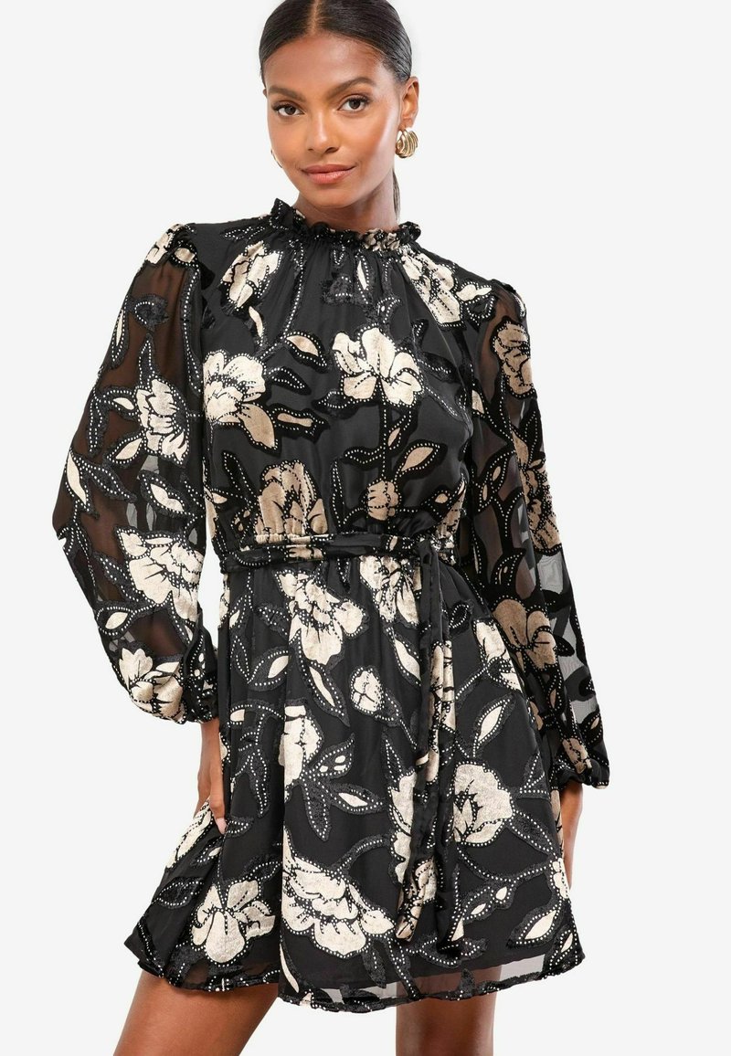 Melnā ziedu kleita ar caurspīdīgām garām piedurknēm, ruffles ap kaklu un saistītu jostasvietu. Detalizētais ziedu raksts ir bēšā un baltā krāsā.